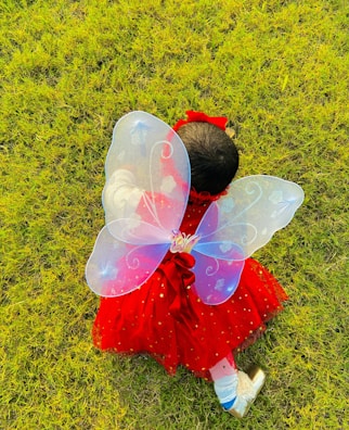 A child wearing a vibrant red dress adorned with gold stars and a large pair of translucent fairy wings crawls or sits on a lush, green grassy field. The wings have a delicate swirl design, and the child is also wearing shiny shoes and a red headband.