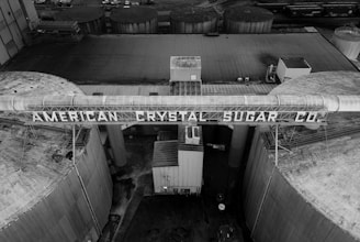 An aerial view of an industrial facility with large cylindrical structures and a prominent sign displaying 'American Crystal Sugar Co.'. The image is in black and white, emphasizing the geometric shapes and textures of the buildings and tanks.