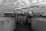 Several large, cylindrical storage tanks dominate the foreground of an industrial plant. Smoke billows from multiple smokestacks, contributing to a dense cloud cover overhead. The atmosphere appears overcast, suggesting a gloomy, industrial setting. The name 'American Crystal Sugar Co.' is prominently displayed on the facility, indicating the nature of the business.