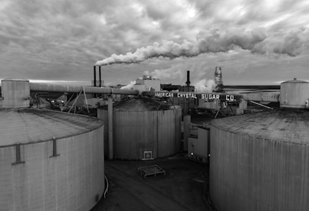 Several large, cylindrical storage tanks dominate the foreground of an industrial plant. Smoke billows from multiple smokestacks, contributing to a dense cloud cover overhead. The atmosphere appears overcast, suggesting a gloomy, industrial setting. The name 'American Crystal Sugar Co.' is prominently displayed on the facility, indicating the nature of the business.