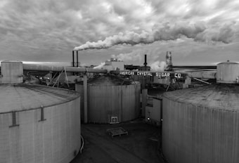 Several large, cylindrical storage tanks dominate the foreground of an industrial plant. Smoke billows from multiple smokestacks, contributing to a dense cloud cover overhead. The atmosphere appears overcast, suggesting a gloomy, industrial setting. The name 'American Crystal Sugar Co.' is prominently displayed on the facility, indicating the nature of the business.