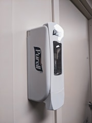 A wall-mounted PURELL hand sanitizer dispenser with a sleek, white design. The dispenser is positioned on a plain, light-colored wall with subtle shadows.