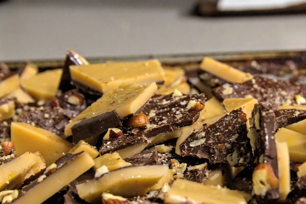 A close-up view of broken pieces of chocolate bark mixed with caramel-colored sections and whole nuts embedded within the bark. The combination of rich dark chocolate and creamy caramel sections is scattered chaotically across a flat surface, creating a textured, rustic appearance.