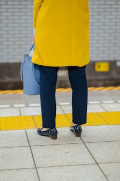 A person dressed in a bright yellow coat is standing on a tiled platform near a tactile paving strip. They are holding a soft blue handbag and wearing dark blue pants and shiny blue shoes. Behind them, a wall with gray bricks is partially visible.