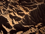 An aerial view of rugged terrain featuring deep, shadowed valleys and sharply defined ridges. The landscape is characterized by its intricate patterns and textures, revealing the natural erosion processes over time.
