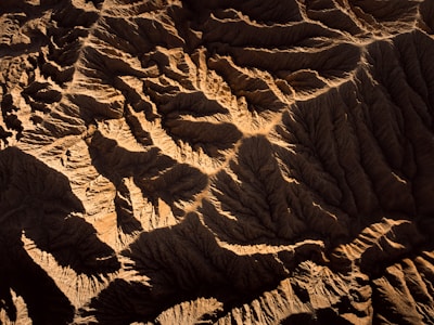 An aerial view of rugged terrain featuring deep, shadowed valleys and sharply defined ridges. The landscape is characterized by its intricate patterns and textures, revealing the natural erosion processes over time.