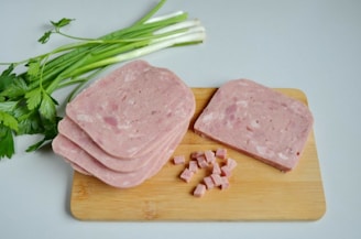 Slices of processed meat are placed on a wooden cutting board next to some finely diced cubes of the same meat. Beside the board, fresh green onions and parsley provide a contrast to the pinkish tones of the meat.