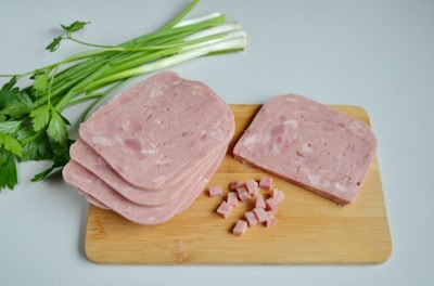 Slices of processed meat are placed on a wooden cutting board next to some finely diced cubes of the same meat. Beside the board, fresh green onions and parsley provide a contrast to the pinkish tones of the meat.