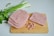 Slices of processed meat are placed on a wooden cutting board next to some finely diced cubes of the same meat. Beside the board, fresh green onions and parsley provide a contrast to the pinkish tones of the meat.