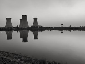 Cooling towers are reflected in the calm water of a large body of water. The image has a monochromatic tone, creating a somber and industrial feel. Vegetation can be seen along the shoreline.