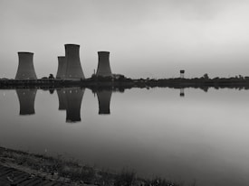 Cooling towers are reflected in the calm water of a large body of water. The image has a monochromatic tone, creating a somber and industrial feel. Vegetation can be seen along the shoreline.
