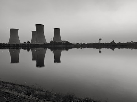 Cooling towers are reflected in the calm water of a large body of water. The image has a monochromatic tone, creating a somber and industrial feel. Vegetation can be seen along the shoreline.