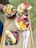 Three acai bowls are garnished with fresh slices of banana, watermelon, orange, strawberries, blueberries, and chocolate chips. The bowls are set on a wooden tray, accompanied by two wooden spoons, suggesting a colorful, healthy, and appetizing presentation.