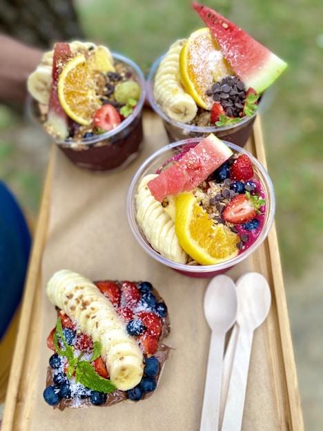 Three acai bowls are garnished with fresh slices of banana, watermelon, orange, strawberries, blueberries, and chocolate chips. The bowls are set on a wooden tray, accompanied by two wooden spoons, suggesting a colorful, healthy, and appetizing presentation.