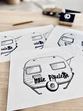 A series of detailed sketches featuring a retro-style camper trailer with the text 'Mlle Pipelette' on each drawing. The sketches are laid out on a light wooden surface, with a pencil and some blurred objects in the background, including a small sign and a wrapped package.