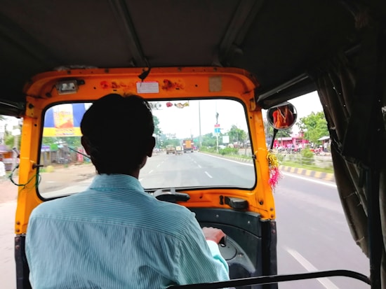 The image captures the interior of a moving auto-rickshaw seen from the passenger's perspective, focusing on the back of the driver's head. The road ahead is visible through the windshield, with various vehicles and roadside buildings visible. The rickshaw's frame is prominent with a decorated interior that includes a string of colorful tassels by the rear-view mirror.