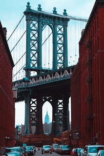 A view of the Manhattan Bridge framed between red brick buildings on either side, with the Empire State Building visible in the distance. Cars are parked along the street, contributing to an urban atmosphere.