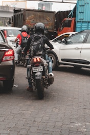 A busy urban street displays a motorcyclist wearing a helmet and backpack, navigating through a blend of stationary cars and large trucks. The scene is set on a paved, cobbled road surrounded by commercial vehicles and city infrastructure.