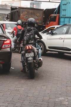 A busy urban street displays a motorcyclist wearing a helmet and backpack, navigating through a blend of stationary cars and large trucks. The scene is set on a paved, cobbled road surrounded by commercial vehicles and city infrastructure.