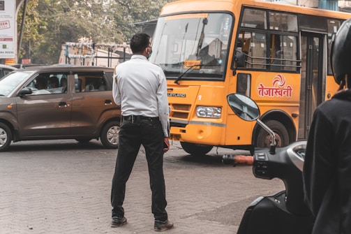 A man in a white shirt standing on a street appears to be monitoring or controlling traffic. A yellow bus with text on it is nearby, facing toward the man, while a brown car waits on the side. Part of a motorcycle with a visible mirror is seen in the foreground. The background includes trees and urban structures.