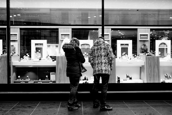 Two people stand in front of a large, brightly lit shop window displaying various luxury items, including jewelry and watches. Both are wearing winter coats, suggesting a cold day.