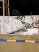 A mural painted on a wall depicting a pair of large shoes stepping onto a small wheelchair, creating a powerful and thought-provoking visual message. The scene is framed by concrete structures and a nighttime backdrop.