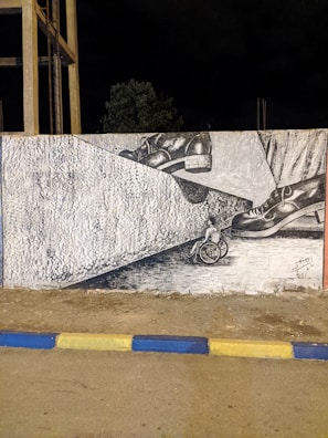 A mural painted on a wall depicting a pair of large shoes stepping onto a small wheelchair, creating a powerful and thought-provoking visual message. The scene is framed by concrete structures and a nighttime backdrop.