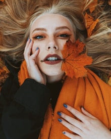 A person with blue nail polish lies on autumn leaves, holding an orange maple leaf near their face. They have light-colored hair spread out around them and wear an orange scarf and dark clothing.