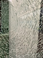 A close-up view of shattered or cracked glass, with intricate patterns of cracks forming across the surface. The background appears indistinct due to the texture of the glass.