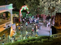 A vibrant nighttime outdoor event with a gathering of people in a park-like setting. Decorative lights are arranged in arches, and there are booths with colorful signs and a leafy tree creating a canopy. The atmosphere is lively, with groups of people interacting and strolling.