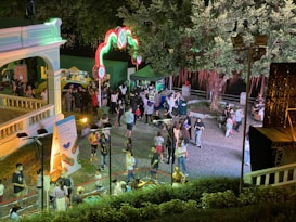 A vibrant nighttime outdoor event with a gathering of people in a park-like setting. Decorative lights are arranged in arches, and there are booths with colorful signs and a leafy tree creating a canopy. The atmosphere is lively, with groups of people interacting and strolling.
