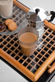 A wooden tray holds a glass of coffee, a stainless steel moka pot, and a small plate with cookies and a chocolate-covered treat. The setting is on a patterned surface, emphasizing a cozy, inviting atmosphere.