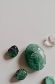 A collection of polished stones displayed on a smooth, light-colored surface. The stones vary in size and shape, with the most prominent being an oval-shaped green stone. Other stones include smaller green and clear stones, each with a shiny, glossy finish.