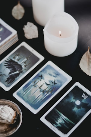 Three tarot cards are laid out on a dark surface alongside a bundle of white sage in a small dish. A white lit candle and several dried leaves are also present. The tarot cards depict various images including swords, birds, and a moonlit scene.