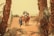 A group of people riding camels is led by a man in traditional attire through a dry, desert landscape, framed by tall palm trees. The sky is hazy, suggesting a hot, arid environment.