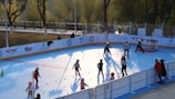 A group of children wearing helmets and roller skates are participating in an outdoor skating activity on a bright blue surface. Some adults are nearby, supervising or instructing the children. The area is enclosed with a barrier, and there are colorful cones set up on the ground. Trees and a pathway are visible in the background, suggesting a park setting.