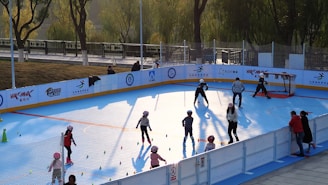 A group of children wearing helmets and roller skates are participating in an outdoor skating activity on a bright blue surface. Some adults are nearby, supervising or instructing the children. The area is enclosed with a barrier, and there are colorful cones set up on the ground. Trees and a pathway are visible in the background, suggesting a park setting.