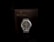A wristwatch with a metal mesh band is placed on a dark brown cushion inside a brown box. The watch face is black with silver hour markers and hands. The words 'Symbol of Uniqueness' are embossed in gold on the box.