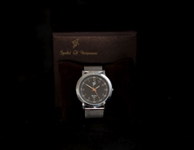 A wristwatch with a metal mesh band is placed on a dark brown cushion inside a brown box. The watch face is black with silver hour markers and hands. The words 'Symbol of Uniqueness' are embossed in gold on the box.