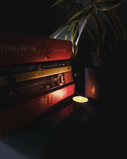 A stack of books is arranged on a dark surface, with titles including 'Harry Potter,' 'The Secret,' and 'My Experiments with Truth.' The dimly lit scene is softened by a small, glowing candle nearby and a green plant draping over from above, casting shadows and adding a cozy atmosphere.
