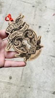 A hand is holding a wooden heart-shaped ornament with intricate black designs and the words '18 Years Strong Feelings Pantheon' etched onto it. There is a red ribbon attached to the top, and the background is a textured concrete surface.