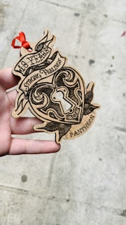 A hand is holding a wooden heart-shaped ornament with intricate black designs and the words '18 Years Strong Feelings Pantheon' etched onto it. There is a red ribbon attached to the top, and the background is a textured concrete surface.