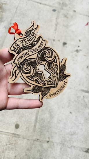 A hand is holding a wooden heart-shaped ornament with intricate black designs and the words '18 Years Strong Feelings Pantheon' etched onto it. There is a red ribbon attached to the top, and the background is a textured concrete surface.