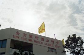A building with red signage in both English and Chinese script reads 'Seow Teck San Physical Cultural Society.' Two yellow flags are visible on the roof, and there are trees in the foreground. The sky appears cloudy with diffuse sunlight.