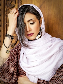 A woman wearing a light hijab, adorned with elegant pearl earrings on her left ear, and a beaded bracelet on her left wrist. She has dark hair and expressive makeup, including bold lipstick and eye makeup. She is touching her hair with her right hand and standing against a wooden carved background.