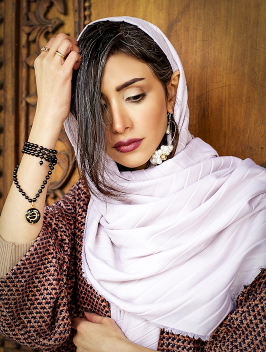 A woman wearing a light hijab, adorned with elegant pearl earrings on her left ear, and a beaded bracelet on her left wrist. She has dark hair and expressive makeup, including bold lipstick and eye makeup. She is touching her hair with her right hand and standing against a wooden carved background.