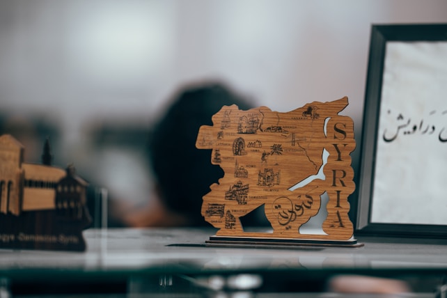 A wooden map of Syria featuring detailed engravings of cities and landmarks is prominently displayed. To the right, a partial view of a framed sign with Arabic script is visible. The background is softly blurred, lending focus to the map.