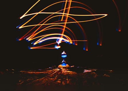 Bright streaks of light in shades of orange and blue create a dynamic pattern against a dark background. At the center, a glowing object, resembling a lamp or bulb, emits a vibrant blue hue. The light patterns loop and swirl above and around this central object.