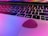 A close-up view of a laptop keyboard with black keys, including a visible Command and Option key. A pink guitar pick rests on the laptop touchpad, and colorful reflections of pink and purple light are present on the surface.