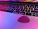 A close-up view of a laptop keyboard with black keys, including a visible Command and Option key. A pink guitar pick rests on the laptop touchpad, and colorful reflections of pink and purple light are present on the surface.